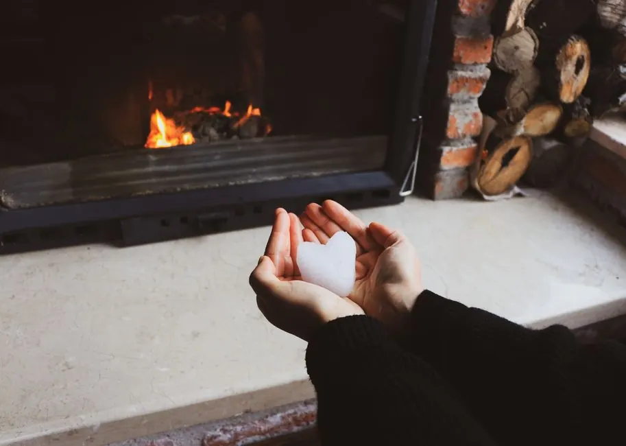 Hands holding heart-shaped snow by a warm fireplace. Cozy winter indoor setting.